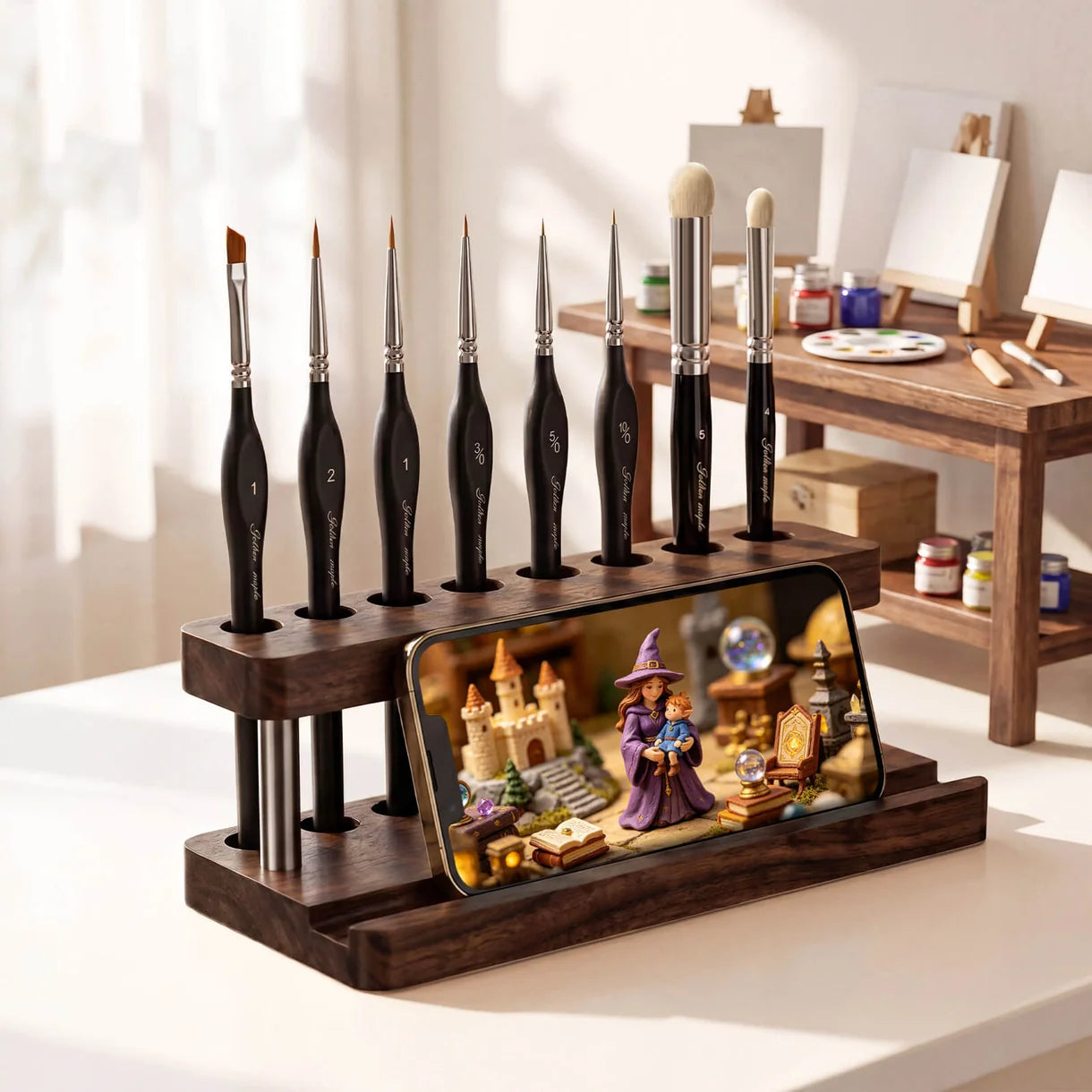 2-in-1 Paint Brush Holder & Phone Stand Golden Maple – lifestyle: drewniany organizer z ciemnego drewna orzechowego z 7 pędzlami Golden Maple (rozmiary 1, 2, 1, 3/0, 3/0, liner, 5) ustawionymi pionowo w otworach górnej belki; w rowku podstawy smartfon wyświetlający scenę fantasy z czarodziejką i zamkiem; w tle miniaturowe biurko malarskie z paletą i farbami