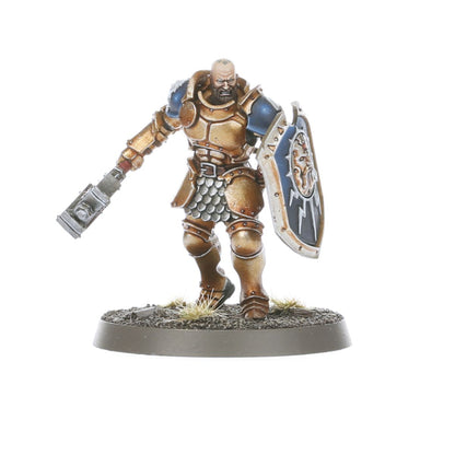 Liberator bez hełmu z brodą - łysy Stormcast Eternal z brodą w złoto-brązowej zbroi, niebieski naramiennik, zamach warhammera w dół, niebieska tarcza z symbolem Sigmara - Warhammer Age of Sigmar