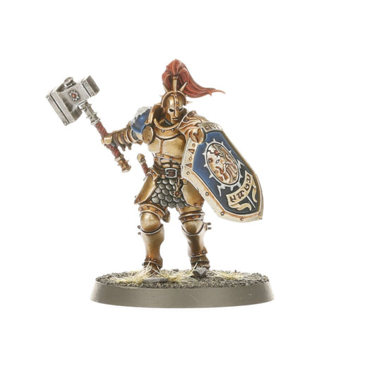 Liberator-Prime z czerwonym pióropuszem - Stormcast Eternal w złotej zbroi z niebieską tarczą z herbem Sigmara i złotym napisem, unosi warhammer w prawej ręce - Warhammer Age of Sigmar