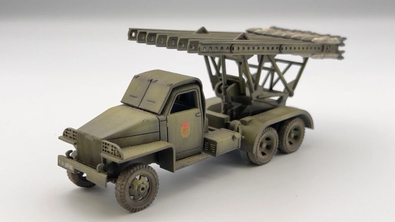 Rubicon Models 280036 BM-13N Katiusza – zmontowany i pomalowany model plastikowy 1/56 28mm widok z boku-przodu: Studebaker US6 6x6 w oliwkowej zieleni z czerwoną gwiazdą na drzwiach, wyrzutnia rakiet M-13 uniesiona pod kątem, szyny startowe z rakietami, koła terenowe 6x6, białe tło, Bolt Action