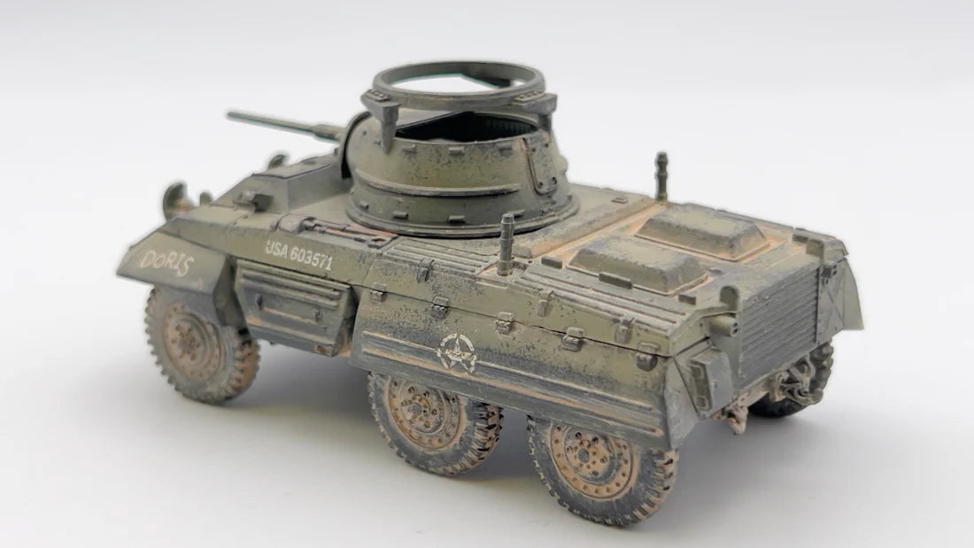 Rubicon Models 280028 M8 Greyhound – zmontowany i pomalowany model plastikowy 1/56 28mm widok z boku: amerykański samochód pancerny w oliwkowej zieleni z napisem DORIS i numerem USA 603571 na kadłubie, armata 37mm M6 w wieży, pierŜień MG, koła terenowe 6x6, napis M8 GREYHOUND, białe tło, Bolt Action