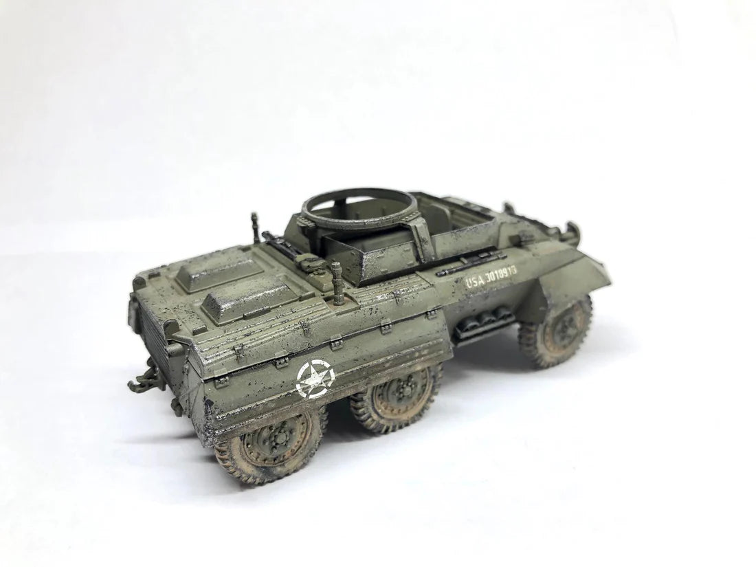 Rubicon Models 280028 M8 Greyhound – zmontowany i pomalowany model plastikowy 1/56 28mm widok z przodu-boku: amerykański samochód pancerny w oliwkowej zieleni z białą gwiazdą i numerem USA 3018913 na kadłubie, pierŜień MG na wieży, właz kierowcy otwarty, koła terenowe 6x6, białe tło, Bolt Action