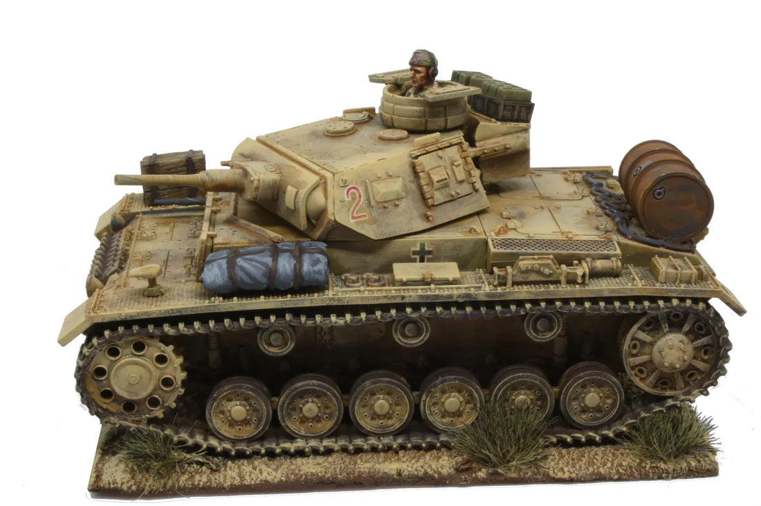 Rubicon Models 280091 Panzer III Ausf. E/F/G – zmontowany i pomalowany model plastikowy 1/56 28mm widok z boku-przodu: PzKpfw III Ausf G w pustynnym kamuflażu piaskowym z numerem 2 i krzyżem żelaznym na wieży, armata 37mm KwK 35/36, figurka dowódcy w otworze wieży, wyposażenie na kadłubie (beczka, bagaż), gąsienice stalowe, białe tło, Bolt Action