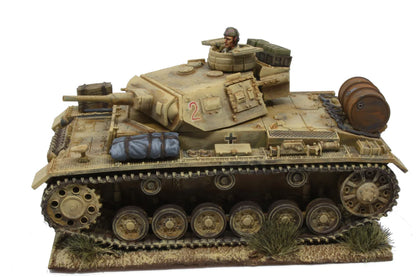 Rubicon Models 280091 Panzer III Ausf. E/F/G – zmontowany i pomalowany model plastikowy 1/56 28mm widok z boku-przodu: PzKpfw III Ausf G w pustynnym kamuflażu piaskowym z numerem 2 i krzyżem żelaznym na wieży, armata 37mm KwK 35/36, figurka dowódcy w otworze wieży, wyposażenie na kadłubie (beczka, bagaż), gąsienice stalowe, białe tło, Bolt Action