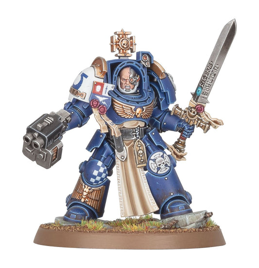 Captain in Terminator Armour Ultramarines - widok z przodu granatowa zbroja terminator złote detale relic sword uniesiony w prawej ręce storm bolter w lewej kremowy tabard cybernetyczne oko brązowa podstawka Warhammer 40K
