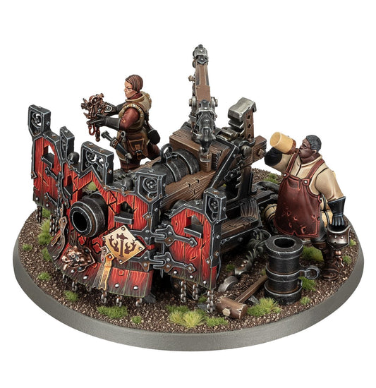 Ironweld Great Cannon - masywna armata Cities of Sigmar na drewnianej platformie z czerwonymi panelami i czarnymi metalowymi okuciami, obsługiwana przez kobietę w brązowym stroju z narzędziami i mężczyznę w czerwonym fartuchu wkładającego ładunek, owalna podstawka z piaskiem i trawą - Warhammer Age of Sigmar