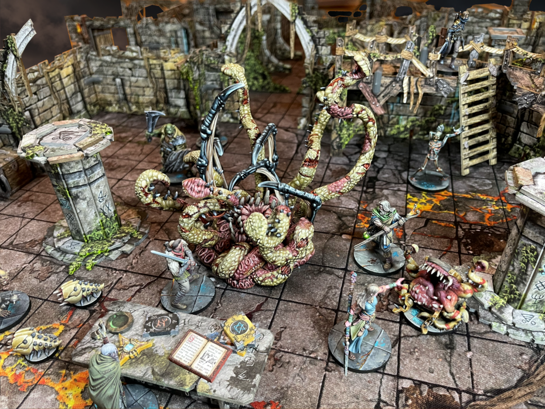 Maladum Oblivion's Maw – The Corrupter Maw z mackami pośrodku areny, Poszukiwacze i Vestige wokół, księga i żetony na pierwszym planie
