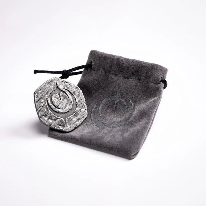 Malediction Metal Initiative Token – awers żetonu z logo Malediction oparty o szary zamszowy woreczek z logo Malediction na jasnym tle