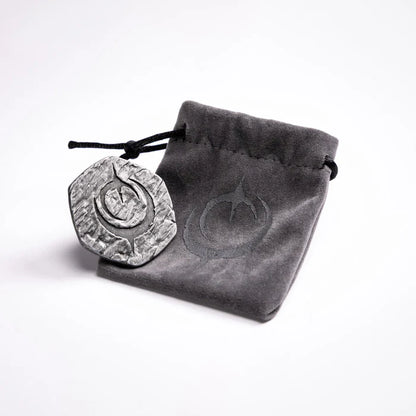 Malediction Metal Initiative Token – awers żetonu z logo Malediction oparty o szary zamszowy woreczek z logo Malediction na jasnym tle