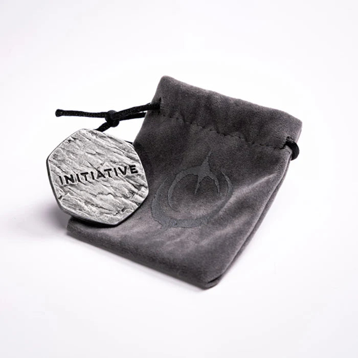 Malediction Metal Initiative Token – rewers żetonu z napisem INITIATIVE oparty o szary zamszowy woreczek z logo Malediction na jasnym tle
