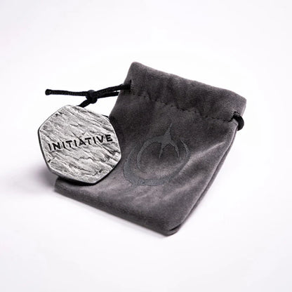 Malediction Metal Initiative Token – rewers żetonu z napisem INITIATIVE oparty o szary zamszowy woreczek z logo Malediction na jasnym tle