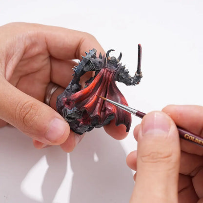 Malowanie figurki demona fantasy pędzlem Golden Maple Master I Kolinsky Sable – precyzyjne detale na czerwonej pelerynie, dłonie trzymające figurkę i pędzel z napisem Golden Maple
