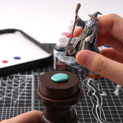 Sticky Putty Golden Maple – lifestyle: dwie dłonie mocujące figurkę czarodzieja (Gandalf) do ciemnobrązowego uchwytu do malowania za pomocą turkusowej masy Sticky Putty; w tle mokra paleta, farby akrylowe i mata do cięcia; zbliżenie na turkusową kulkę masy na górze uchwytu
