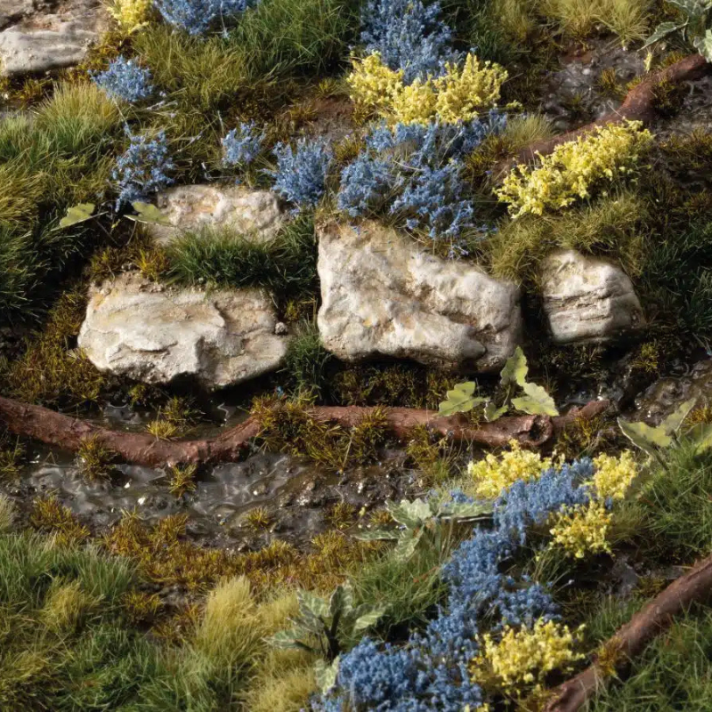 Bluebeard Flowers 6mm Gamers Grass - diorama leśna z kamieniami, korzeniami i strumieniem pokryta niebieskimi i żółtymi kępkami kwiatowymi wśród zielonej trawy

