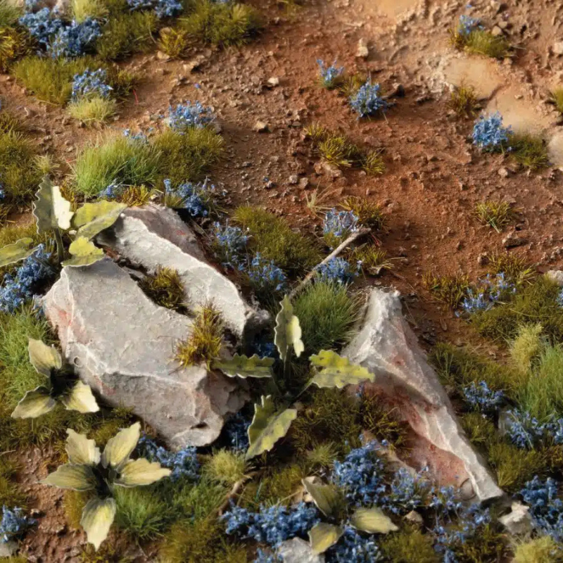 Cornflower Tiny Flowers 4mm Gamers Grass - diorama z czerwoną ziemią i kamieniami pokryta drobnymi niebieskimi kępkami chabra wśród zielonej trawy i roślin
