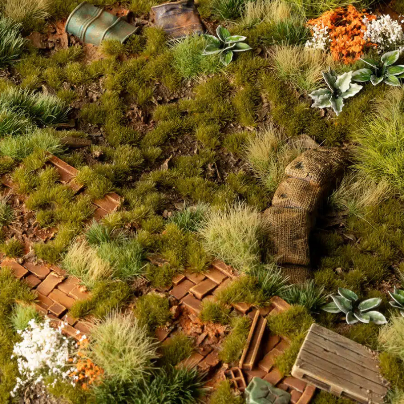 Dry Green Tufts 2mm Wild Gamers Grass - diorama z cegłami, workami i beczkami pokryta gęstymi drobnymi kępkami suchej zielonej trawy tworzącymi realistyczny, zaniedbany teren
