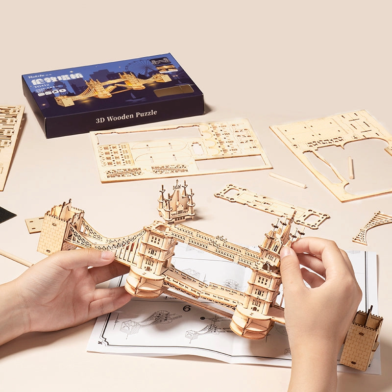 Drewniany puzzle 3D Tower Bridge Rolife RA003e – etap składania: dwie ręce trzymające model, obok pudełko Rolife 3D Wooden Puzzle i arkusze elementów na beżowym tle
