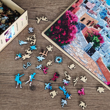 Drewniane puzzle Santorini Bliss Värvikas PW048e – rozsypane figurkowe elementy na drewnianym stole obok częściowo ułożonego obrazu: tancerz, trójząb, słońce, kwiaty i inne kształty
