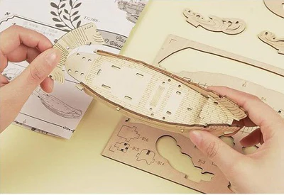 Drewniany puzzle 3D Żaglowiec Rolife RA005e – etap składania: ręce trzymające częściowo złożony kadłub statku, obok arkusze ze sklejki i instrukcja
