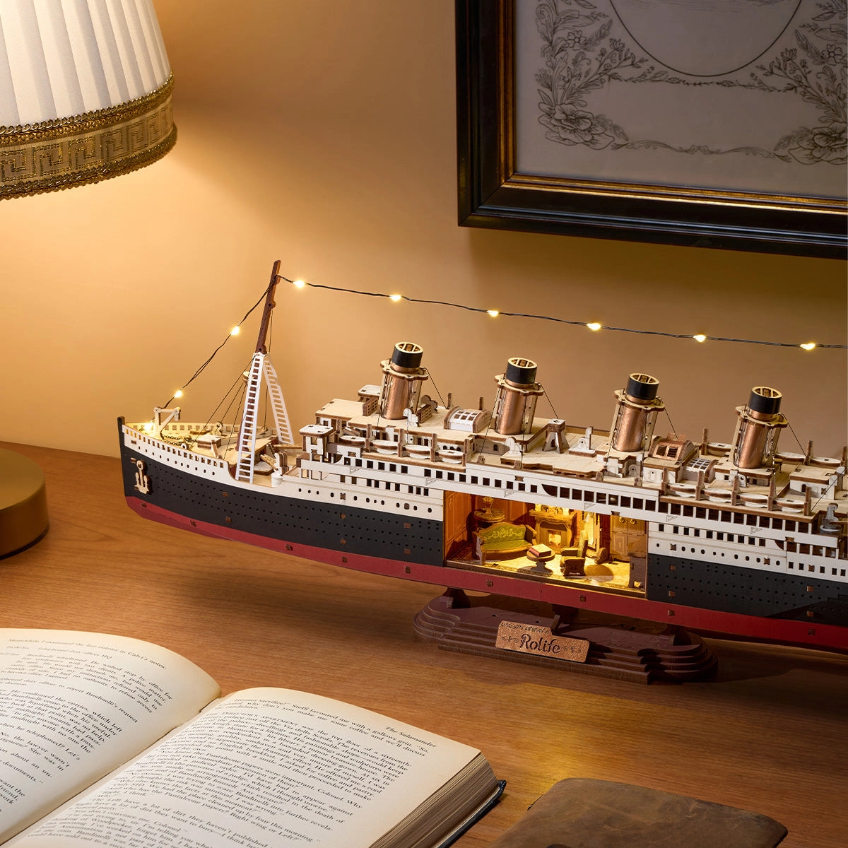 Drewniany puzzle 3D 1912 Eternal Atlantic Voyager Rolife TGL02 – ukończony model na drewnianym stole z podświetlonym wnętrzem i światłami LED na olinowaniu, w tle obraz i lampa, książka
