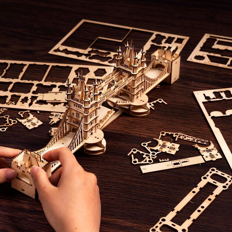 Drewniany puzzle 3D Tower Bridge Rolife RA003e – etap składania: ręka trzymająca częściowo złożoną wieżę mostu, obok arkusze ze sklejki na ciemnym stole
