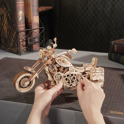 Mechaniczny drewniany model 3D Motocykl Cruiser Rolife RK026e – ręce trzymające prawie ukończony model na ciemnym stole, w tle książki i dekoracje
