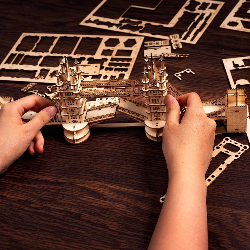 Drewniany puzzle 3D Tower Bridge Rolife RA003e – etap składania: dwie ręce trzymające prawie ukończony most, widoczne obie wieże i łącząca je kratownica
