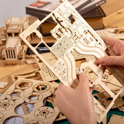 Mechaniczny drewniany model 3D Samochód Grand Prix V8 Rolife RA001e – ręce trzymające arkusz sklejki z wyciętymi elementami, w tle inne modele i książki
