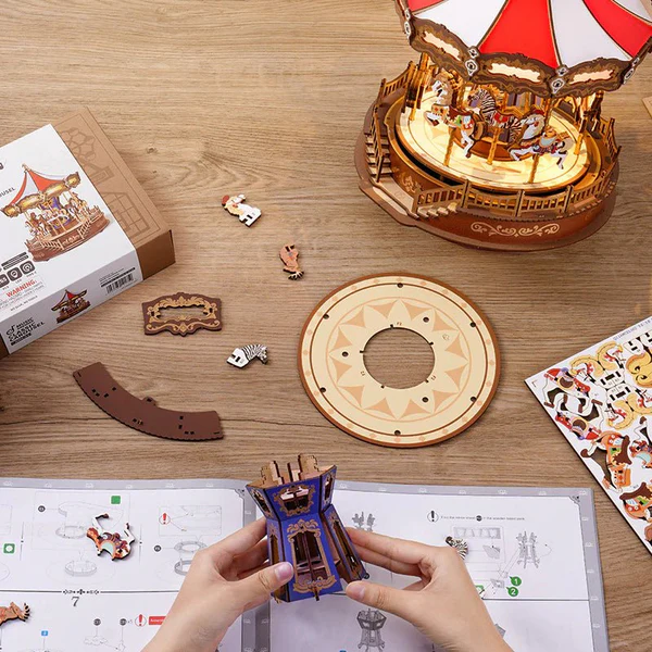 Drewniany puzzle 3D Klasyczna Karuzela Rolife AMC01e – etap składania: ręce trzymające częściowo złożoną podstawę, obok arkusze z elementami, naklejki i instrukcja
