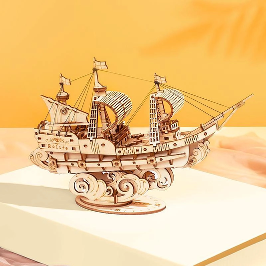 Drewniany puzzle 3D Żaglowiec Rolife RA005e – ukończony model na książce na żółtym tle, widok z boku z żaglami i ozdobnymi falami podstawy

