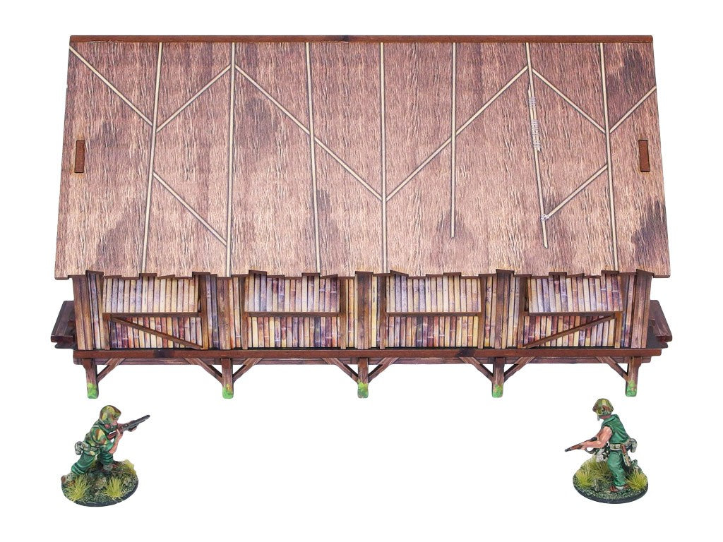 WW2 Pacific Barracks (1) PREPAINTED – zmontowane koszary dżunglowe na palach, widok z przodu: dach ze strzechy z bambusowymi ścianami i werandą, dwie figurki żołnierzy 28mm Bolt Action, białe tło
