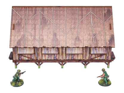 WW2 Pacific Barracks (1) PREPAINTED – zmontowane koszary dżunglowe na palach, widok z przodu: dach ze strzechy z bambusowymi ścianami i werandą, dwie figurki żołnierzy 28mm Bolt Action, białe tło

