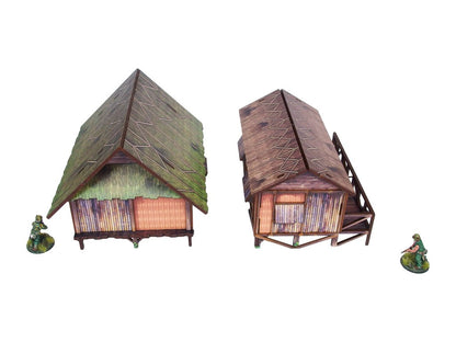 WW2 Pacific Jungle Huts (2) PREPAINTED – dwie zmontowane chaty dżunglowe na palach, widok z przodu pod kątem: zielona strzecha (lewa) i brązowa strzecha z werandą (prawa), figurki żołnierzy 28mm Bolt Action, białe tło
