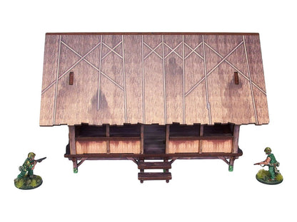 WW2 Pacific Kitchen (1) PREPAINTED – zmontowana kuchnia dżunglowa na palach, widok z przodu: otwarty budynek z dachem ze strzechy w rombowy wzór i bambusowymi ścianami, dwie figurki żołnierzy 28mm Bolt Action, białe tło
